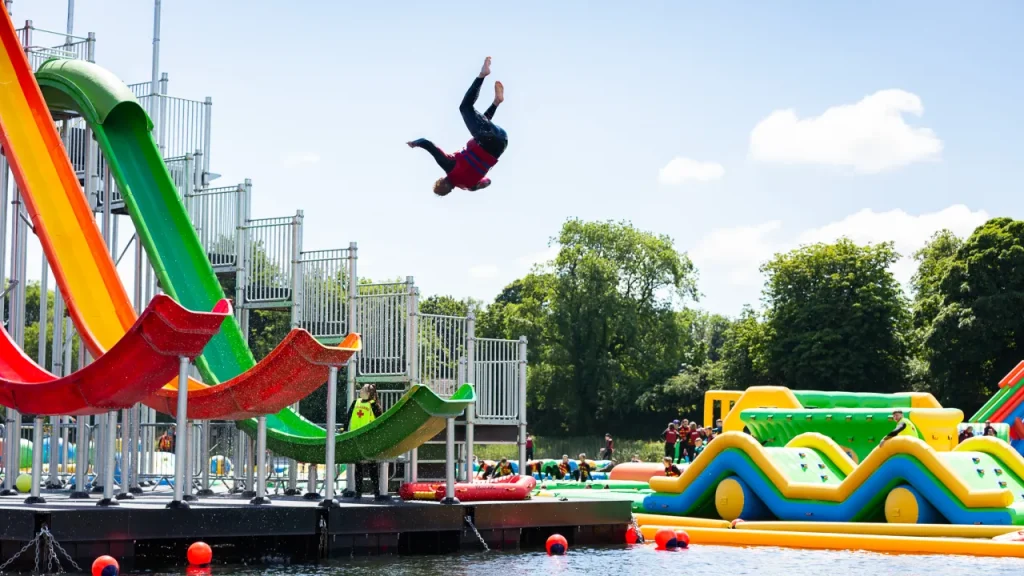 Rider performing a backflip off the green launch slide at The Lake Kilrea waterpark, with multi-lane slides and inflatable obstacles on the lake in Kilrea, County Londonderry.