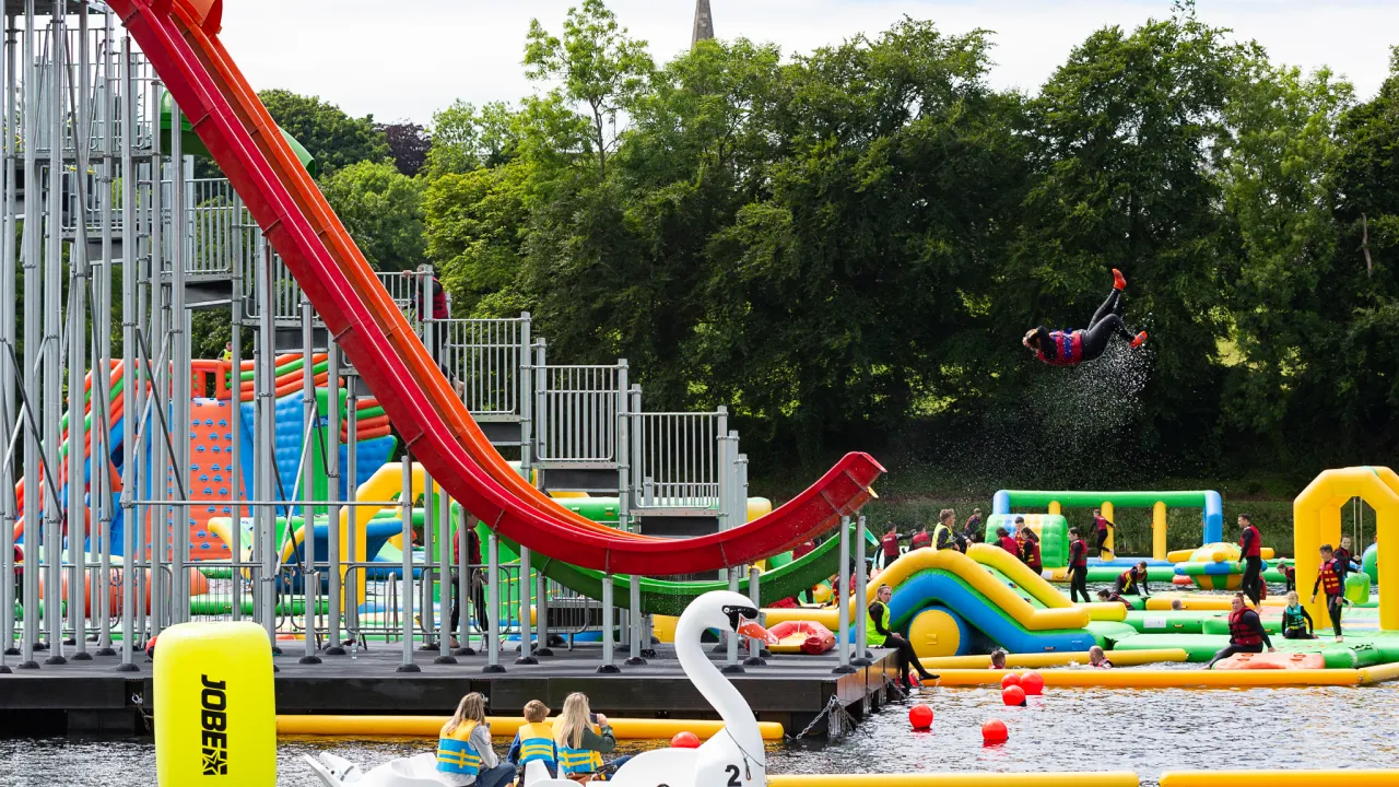 Participant mid-air flipping from a red curved launch slide at The Lake Kilrea waterpark, with multi-lane slides, inflatable obstacles, a swan pedalo and spectators on the spring-fed lake in Kilrea, County Londonderry.