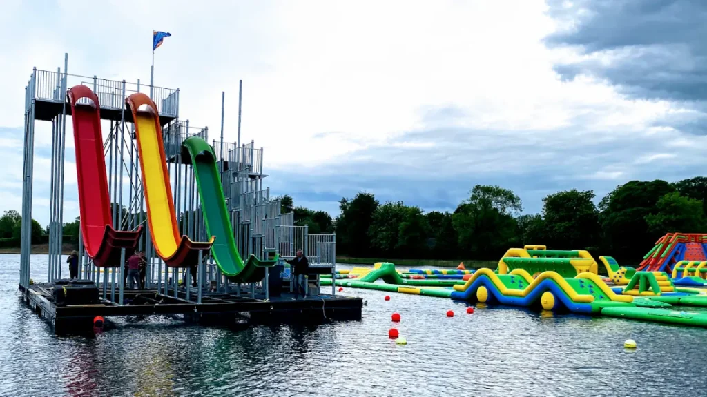 Tall red, yellow, and green slides beside inflatable obstacle courses at The Lake Kilrea Waterpark on a cloudy day with trees in the background.