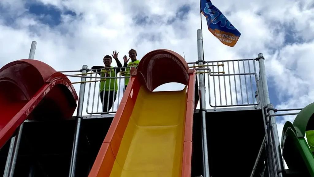 Two lifeguards standing at the top of a tall yellow slide at The Lake Kilrea Waterpark, waving under a partly cloudy sky with the park flag flying.