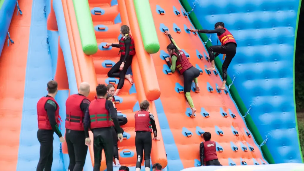 Visitors wearing red life jackets climbing a large orange and blue inflatable wall at The Lake Kilrea Waterpark, surrounded by other guests waiting their turn.