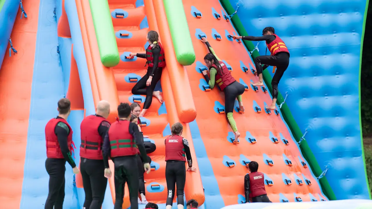 Visitors wearing red life jackets climbing a large orange and blue inflatable wall at The Lake Kilrea Waterpark, surrounded by other guests waiting their turn.