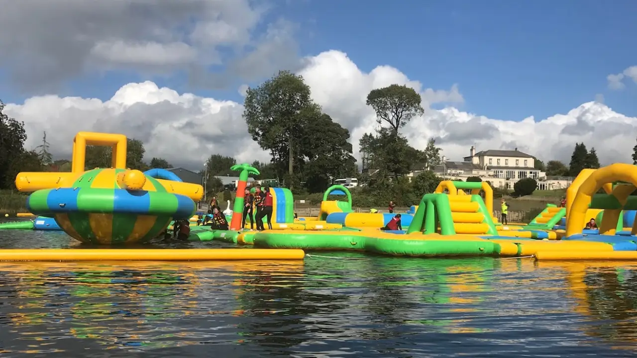 Colourful green and yellow inflatable obstacle course with palm tree decorations at The Lake Kilrea Waterpark, with families enjoying activities under a sunny sky.