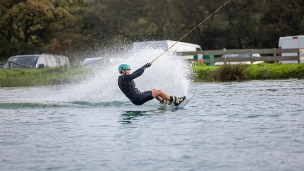 Wakeboarder in helmet and wetsuit carving across the lake at Wild Lakes Aqua Park, with a powerful water spray and greenery in the background.