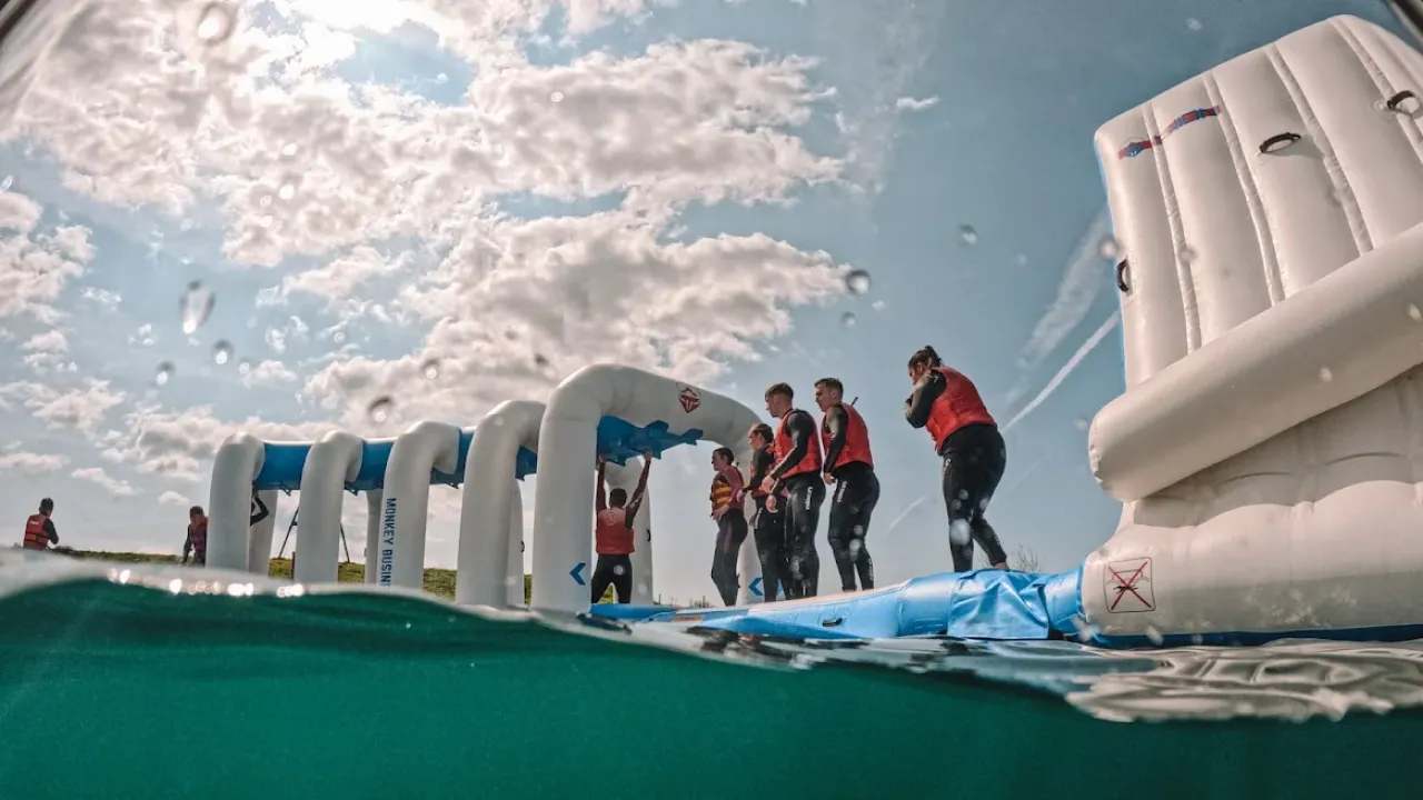 Group of people in wetsuits and life jackets climbing inflatable monkey bars at Wild Lakes Aqua Park in Wales, with half-submerged water view.