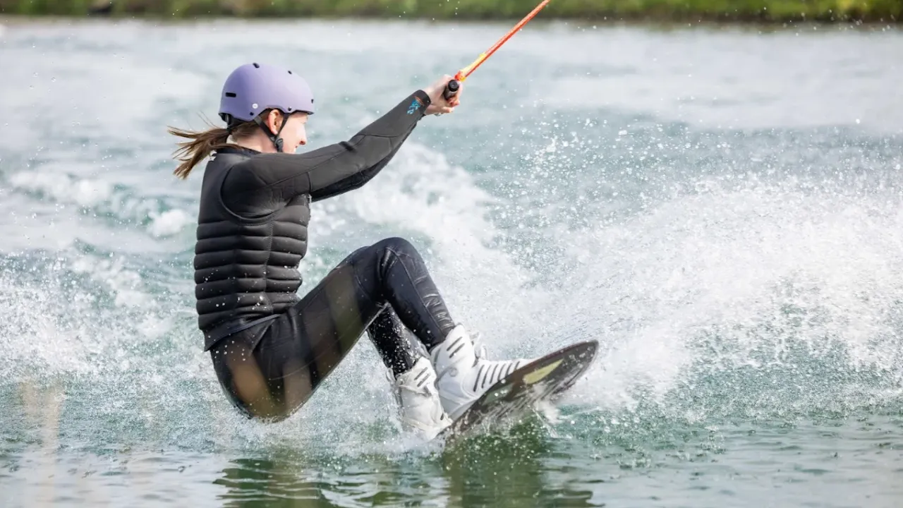 Young woman wakeboarding at Wild Lakes Aqua Park in Wales, wearing a helmet and wetsuit, creating a splash across the lake surface.