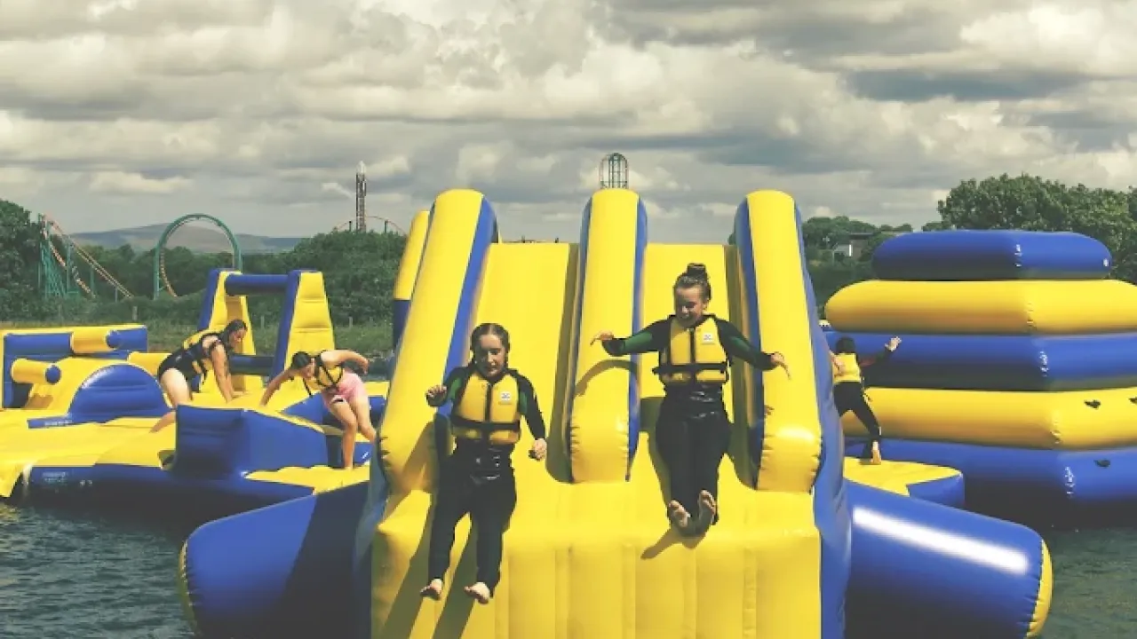 Children in life jackets jumping from yellow and blue inflatable slides at Wild Lakes Aqua Park in Wales on a sunny day, with rollercoasters in background.
