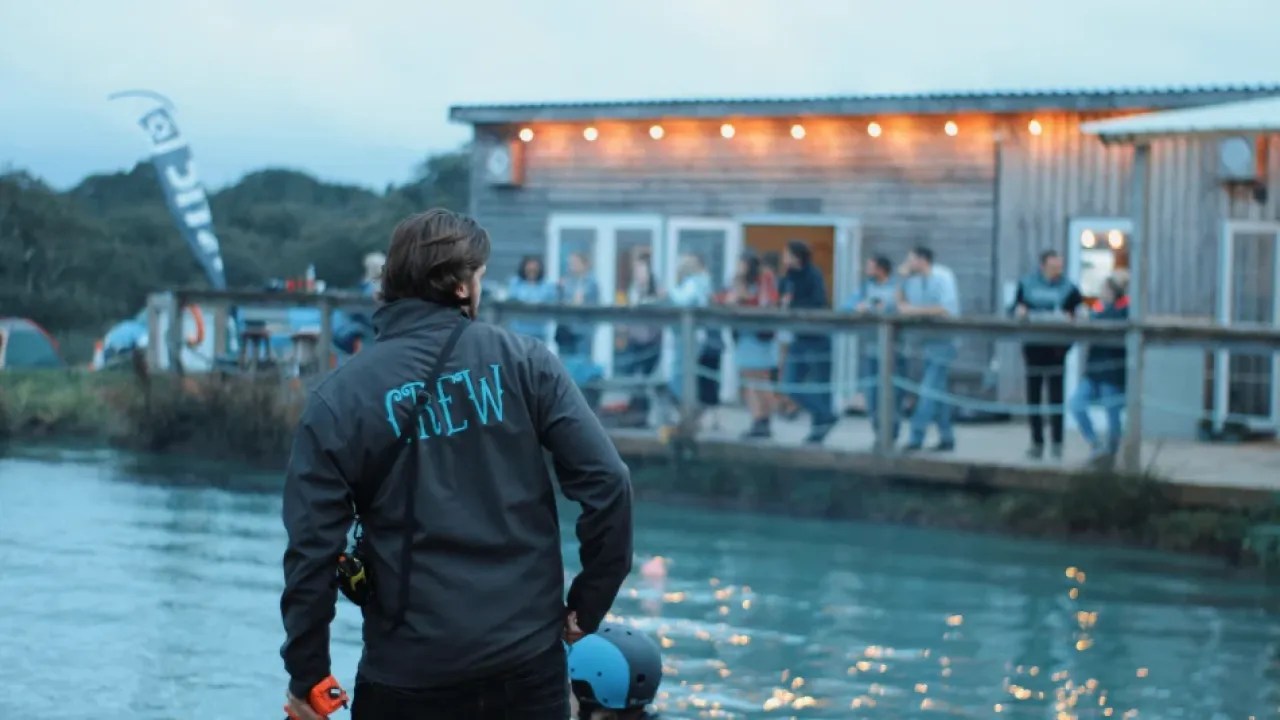 Wild Lakes Aqua Park crew member by the lakeside with people gathered on a wooden deck at dusk near the restaurant in Pembrokeshire, Wales.