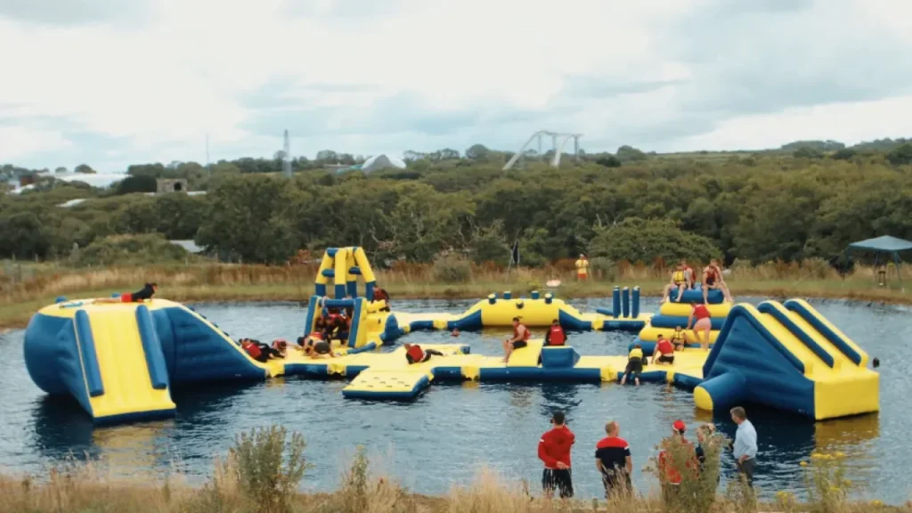 View of guests playing on the large yellow and blue inflatable obstacle course at Wild Lakes Aqua Park, with forest and rollercoasters in the background.