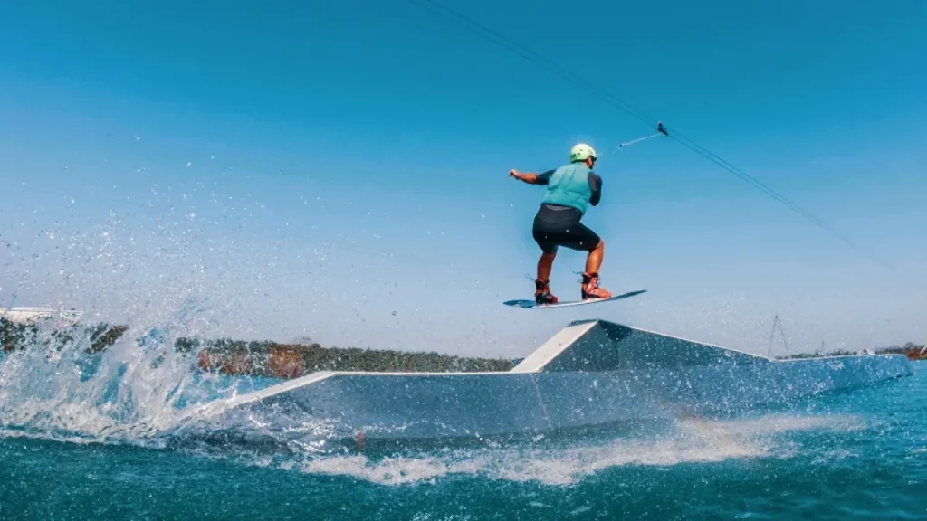 Wakeboarder launching off a ramp at Wild Lakes Aqua Park in Wales, mid-air with splash trails under a clear blue sky.