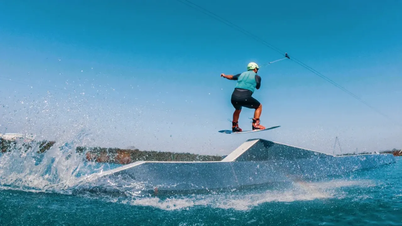 Wakeboarder launching off a ramp at Wild Lakes Aqua Park in Wales, mid-air with splash trails under a clear blue sky.
