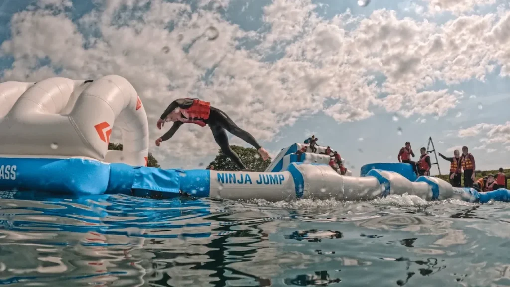 Person mid-flip on the Ninja Jump inflatable obstacle at Wild Lakes Aqua Park, with others watching and cheering in the background on a sunny day.