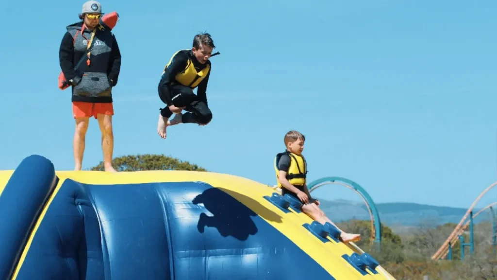 Child mid-air jumping from an inflatable at Wild Lakes Aqua Park while another child and a lifeguard look on, with clear skies and rollercoasters behind.