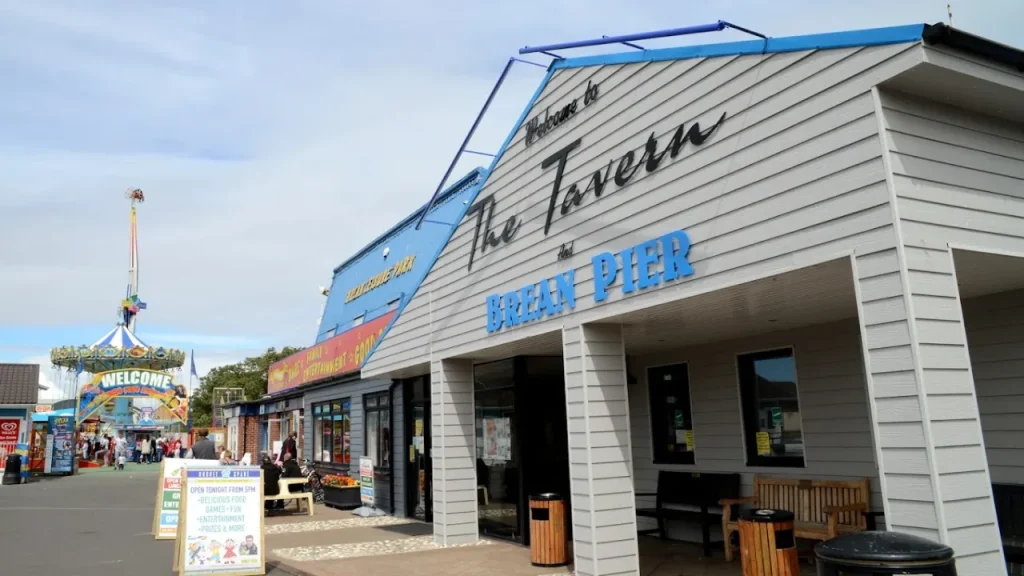Front of The Tavern and Brean Pier entertainment complex at Brean Leisure Park, with signs, seating, and nearby amusement rides.