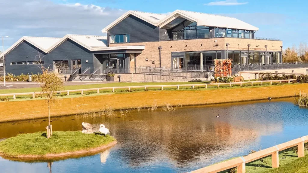 Modern building of Brean Country Club reflected in a lake, with swans on a grassy island and landscaped surroundings near Brean Leisure Park.