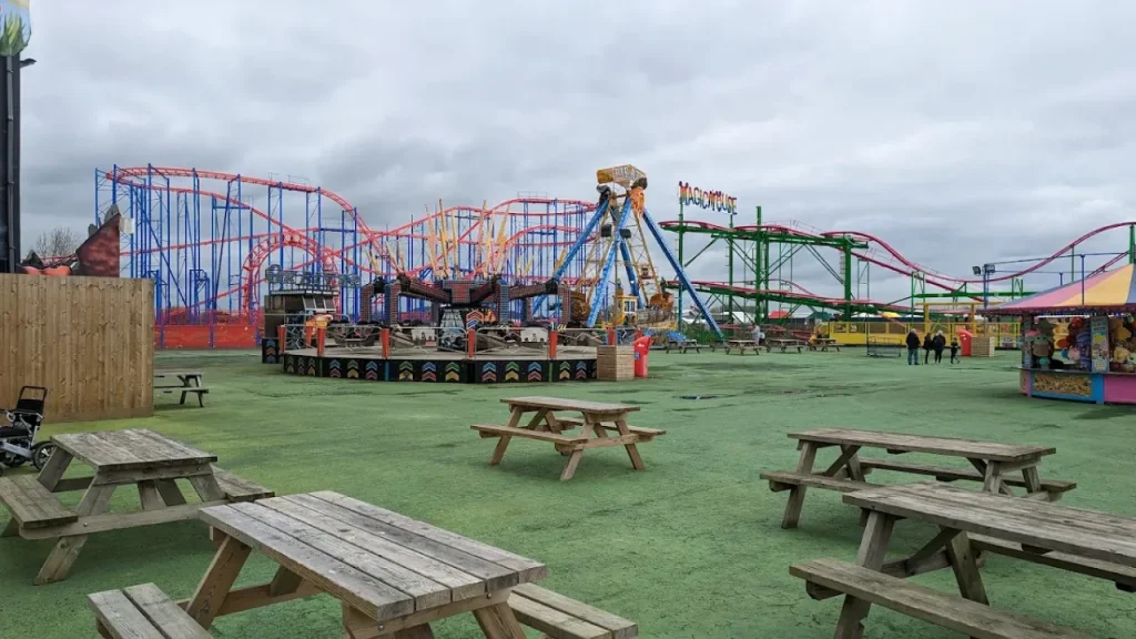 Wooden picnic tables in the open seating area at Brean Leisure Park, with rollercoasters and thrill rides visible in the background.