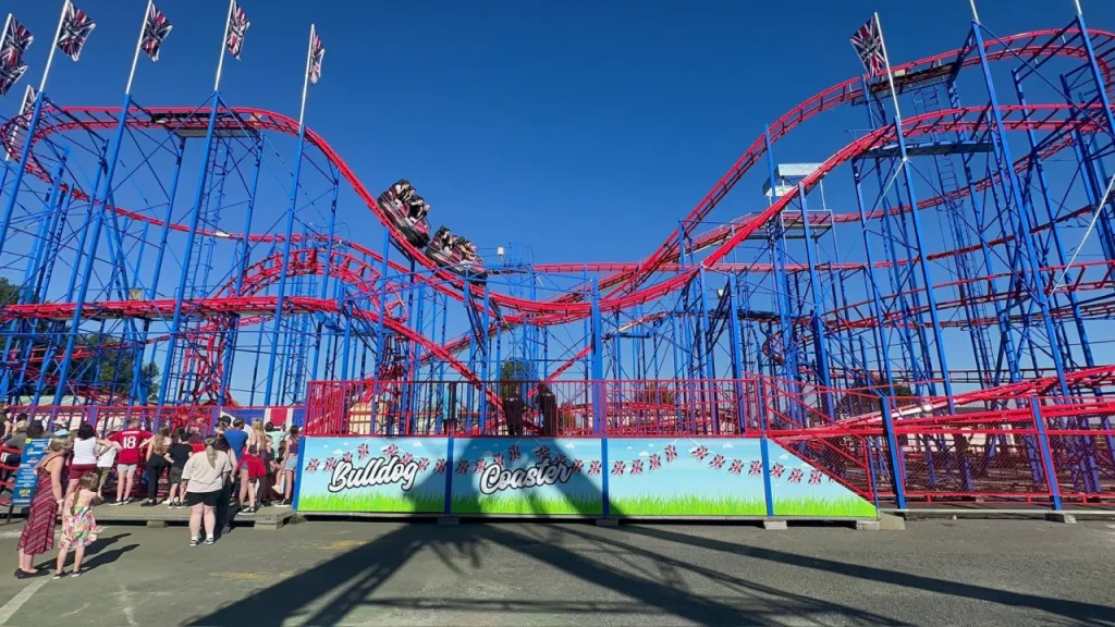 Visitors queue for the Bulldog Coaster, a red and blue looping rollercoaster at Brean Theme Park in Somerset, under a clear blue sky.