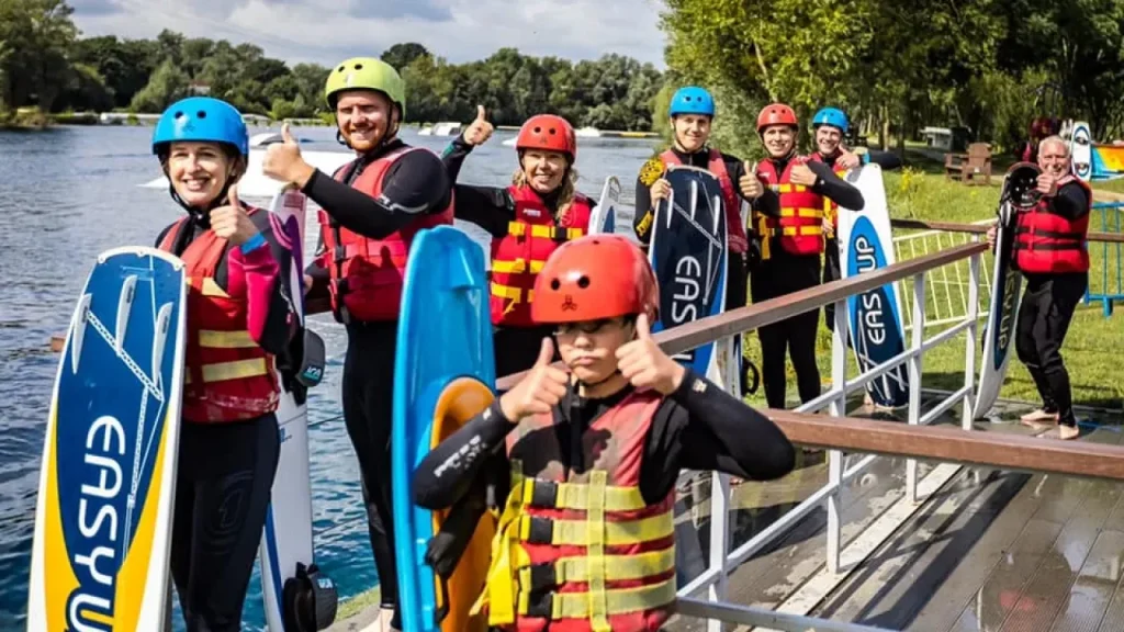 Group of adults and children in wetsuits and helmets holding boards at Liquid Leisure’s wakeboarding dock.