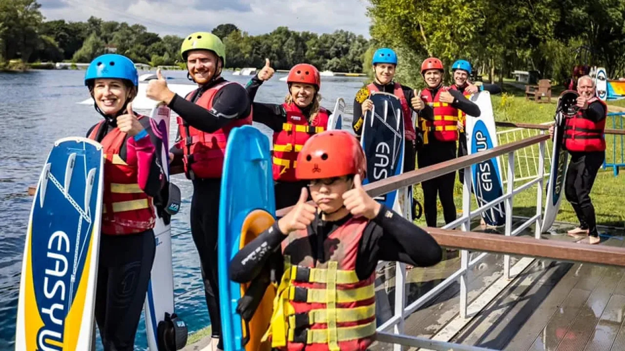 Group of adults and children in wetsuits and helmets holding boards at Liquid Leisure’s wakeboarding dock.
