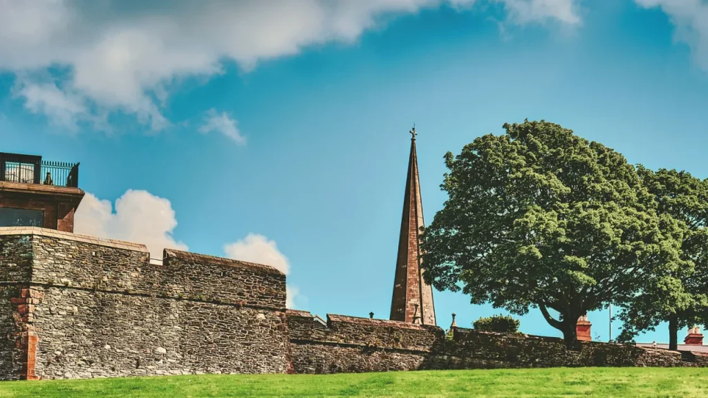 Stone walls of Londonderry city with a Celtic cross and church spire under blue skies in County Londonderry, Northern Ireland.
