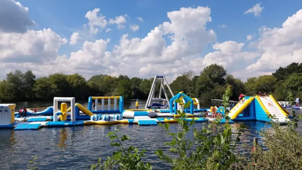 Wide view of the floating inflatable water park at Thorpe Lakes Aqua Park with slides and obstacle structures on a lake under a blue sky.