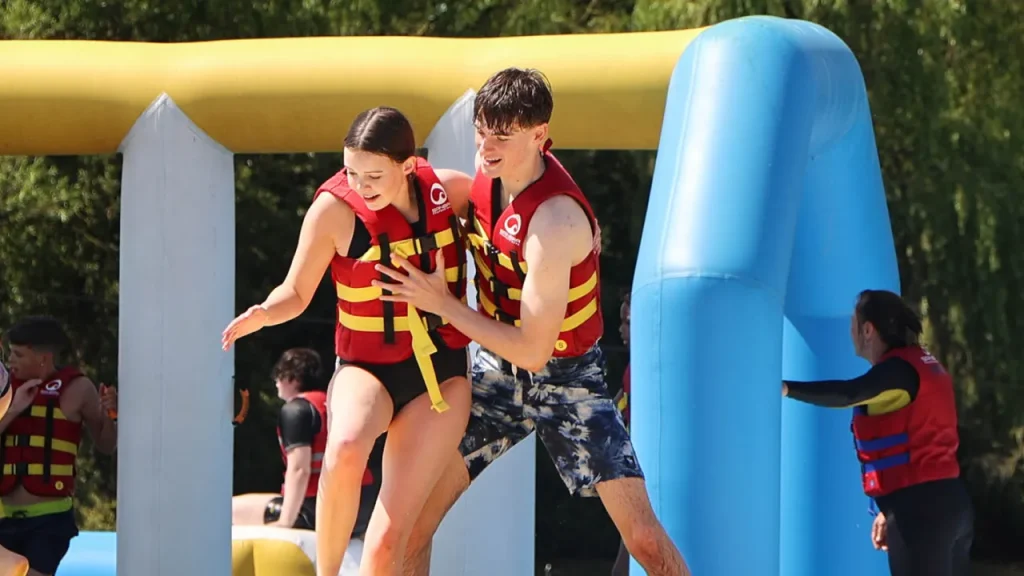 Young couple on an inflatable obstacle at Thorpe Lakes Aqua Park, both wearing red life jackets and enjoying the water activity.