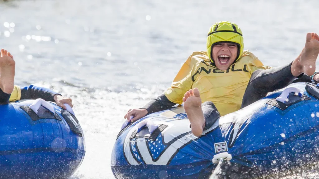 Smiling child in a yellow helmet and wetsuit riding an inflatable tube across the water at Thorpe Lakes Aqua Park.