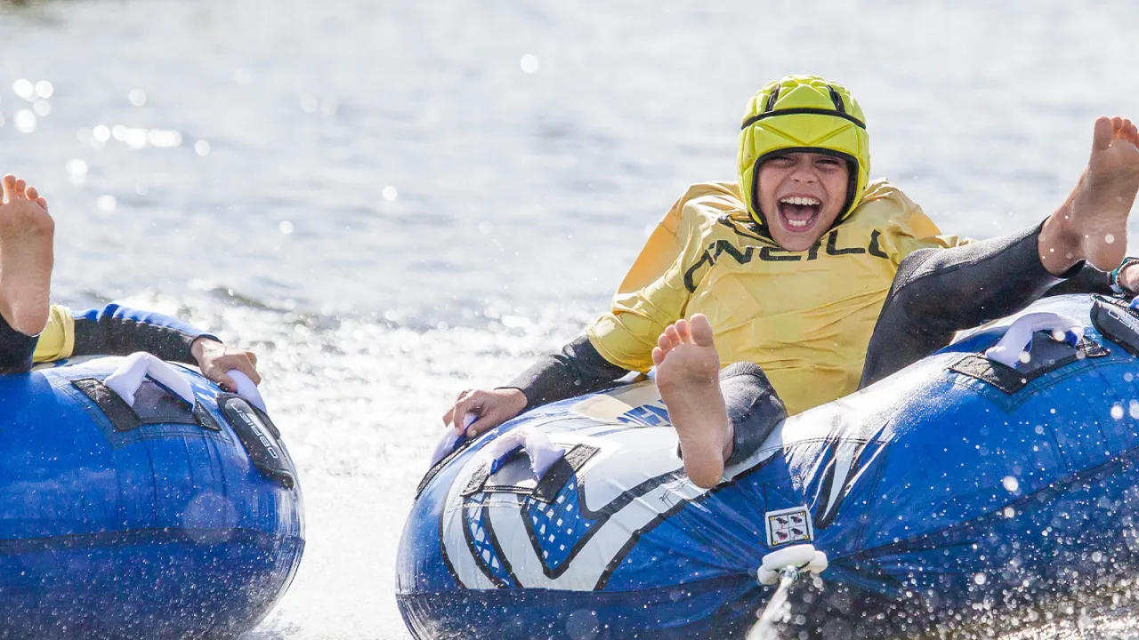 Smiling child in a yellow helmet and wetsuit riding an inflatable tube across the water at Thorpe Lakes Aqua Park.