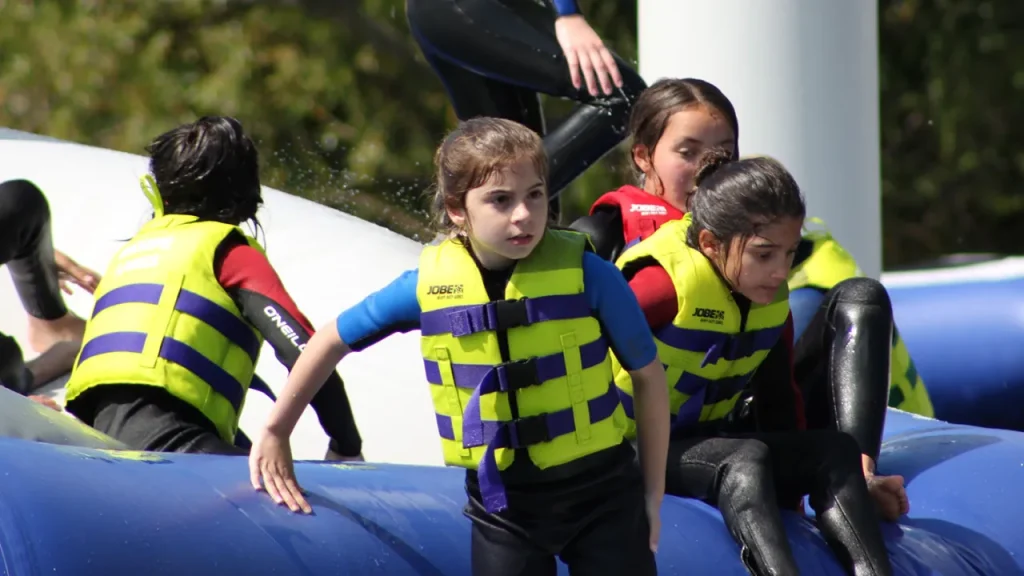 Group of children in wetsuits and yellow-green life jackets playing on inflatable water obstacles at Thorpe Lakes Aqua Park.