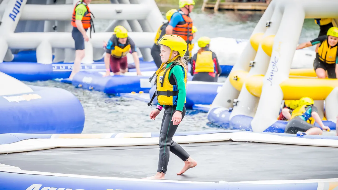 Child in wetsuit and helmet walking across inflatable water equipment at Thorpe Lakes Aqua Park surrounded by other participants.