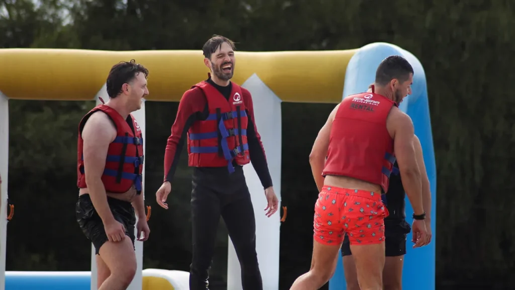 Adult men smiling and playing on the floating inflatable course at Thorpe Lakes Aqua Park, wearing red safety vests.