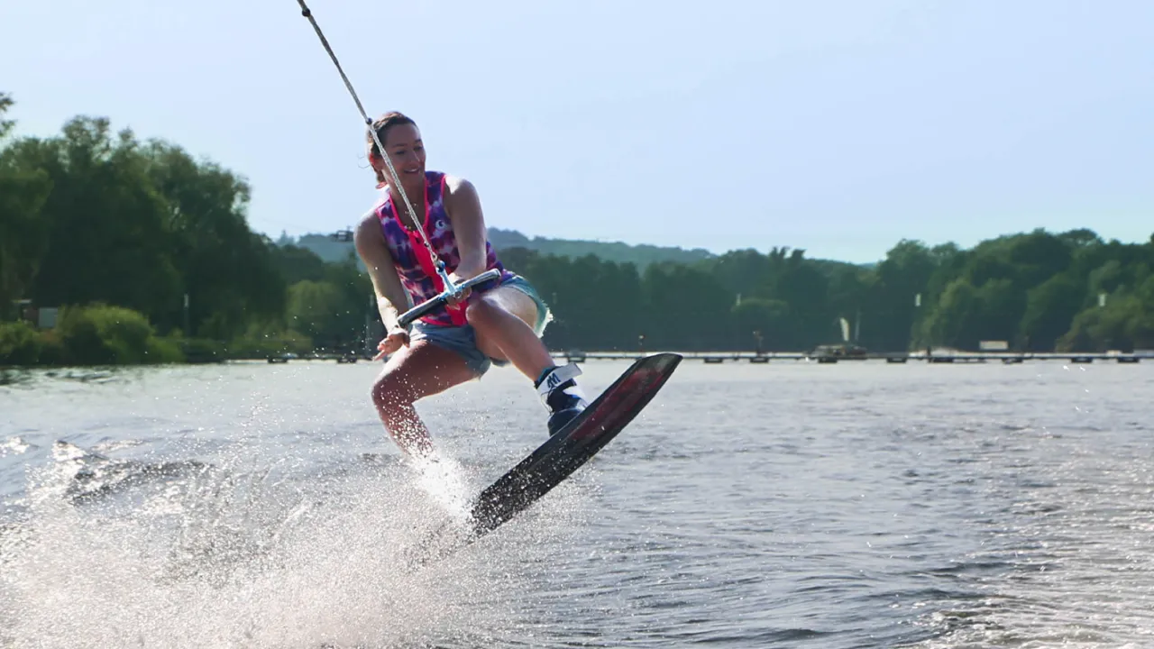 Female wakeboarder mid-air performing a jump on the lake at Thorpe Lakes, with green trees and water in the background.
