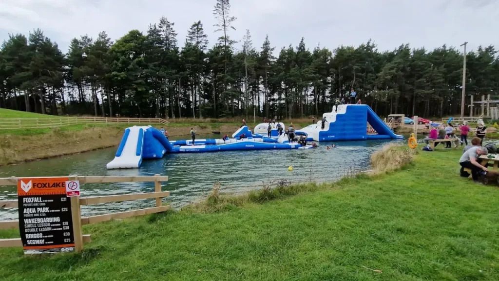 View of the inflatable Aqua Park at Foxlake Adventures in Dunbar, Scotland, with visitors playing on water obstacles and families relaxing on the grassy shore.