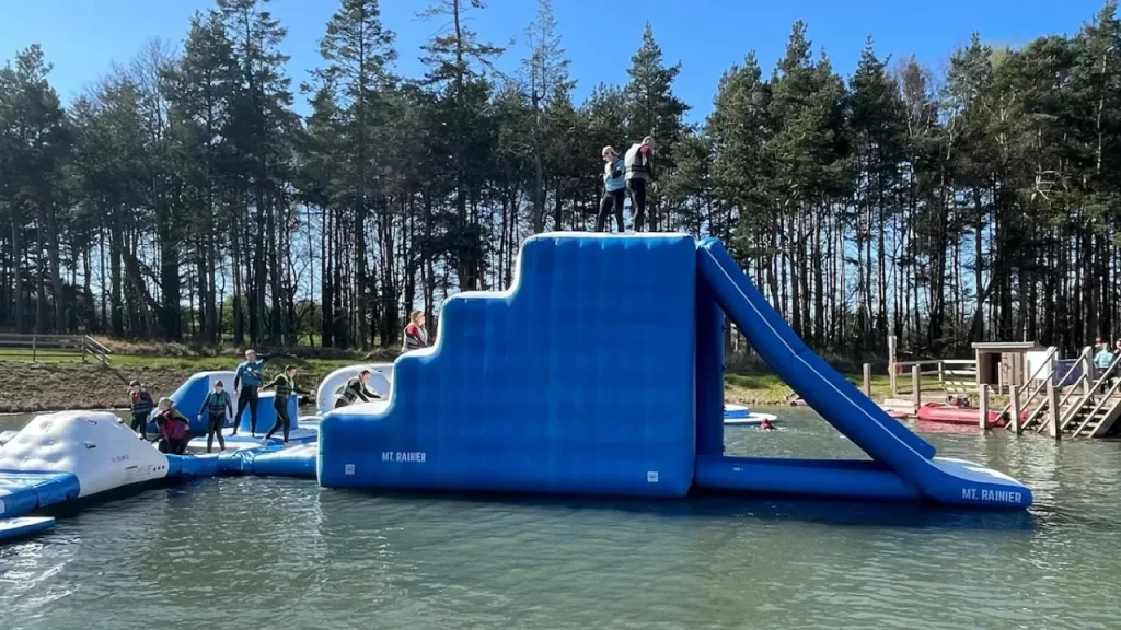 People climbing and sliding down the large blue inflatable “Mt. Rainier” structure at Foxlake Aqua Park, surrounded by forest and open water.