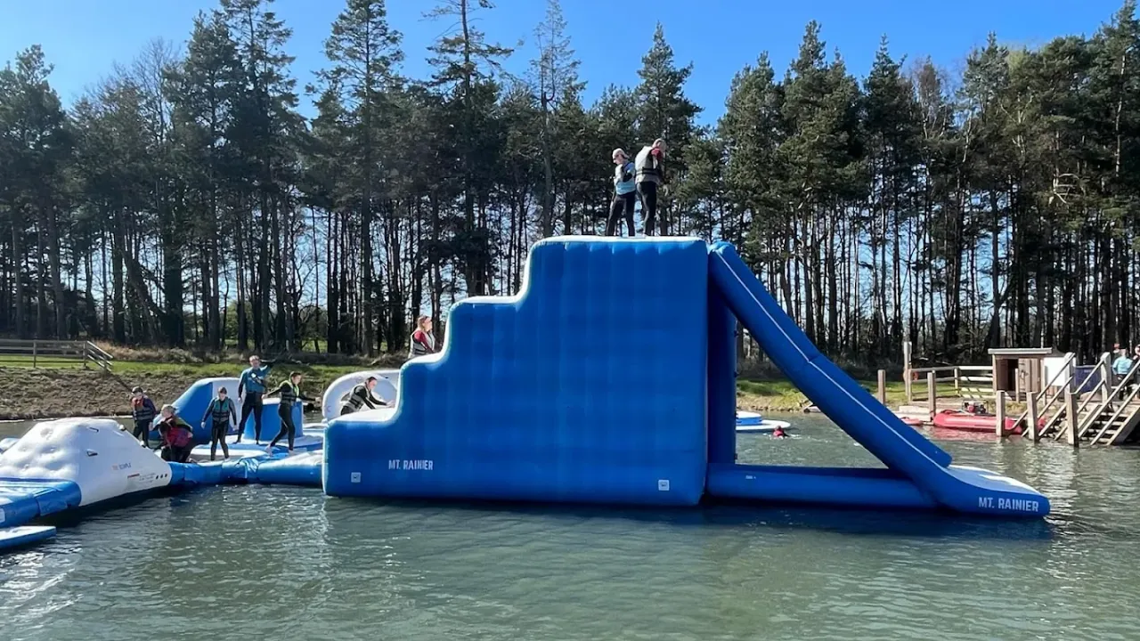 People climbing and sliding down the large blue inflatable “Mt. Rainier” structure at Foxlake Aqua Park, surrounded by forest and open water.