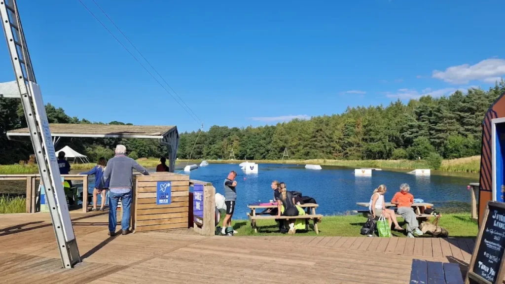 People relaxing at the lakeside dock at Foxlake Adventures Dunbar, with cable wakeboarding equipment and obstacles visible on the water.