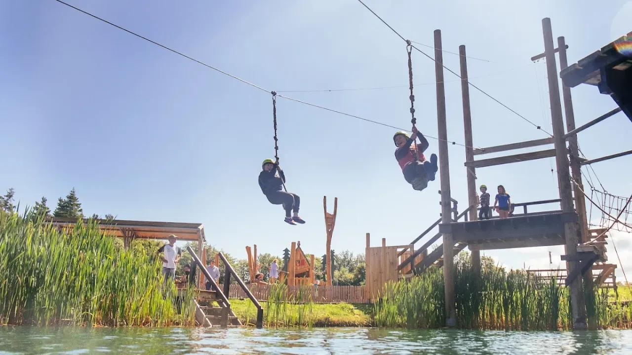 Two children ziplining across a water obstacle course at Foxlake Adventures, wearing helmets and harnesses under clear blue skies.