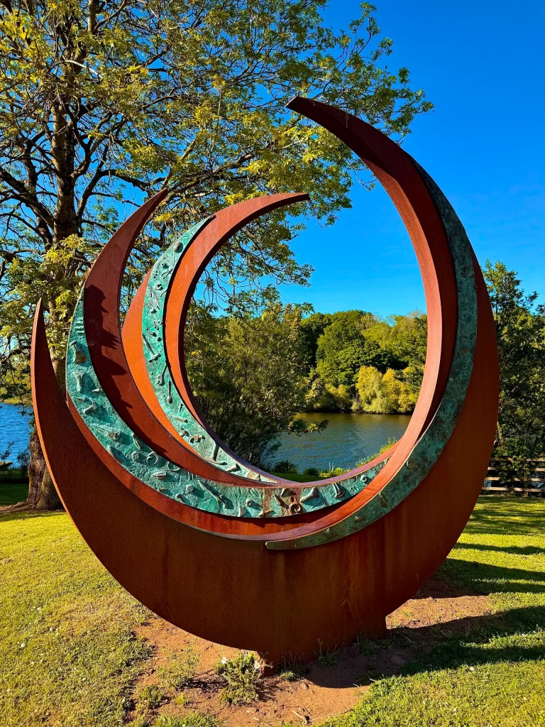 Modern crescent-shaped metal sculpture in Riverside Park, Ballymoney, with a backdrop of trees and the River Bann.