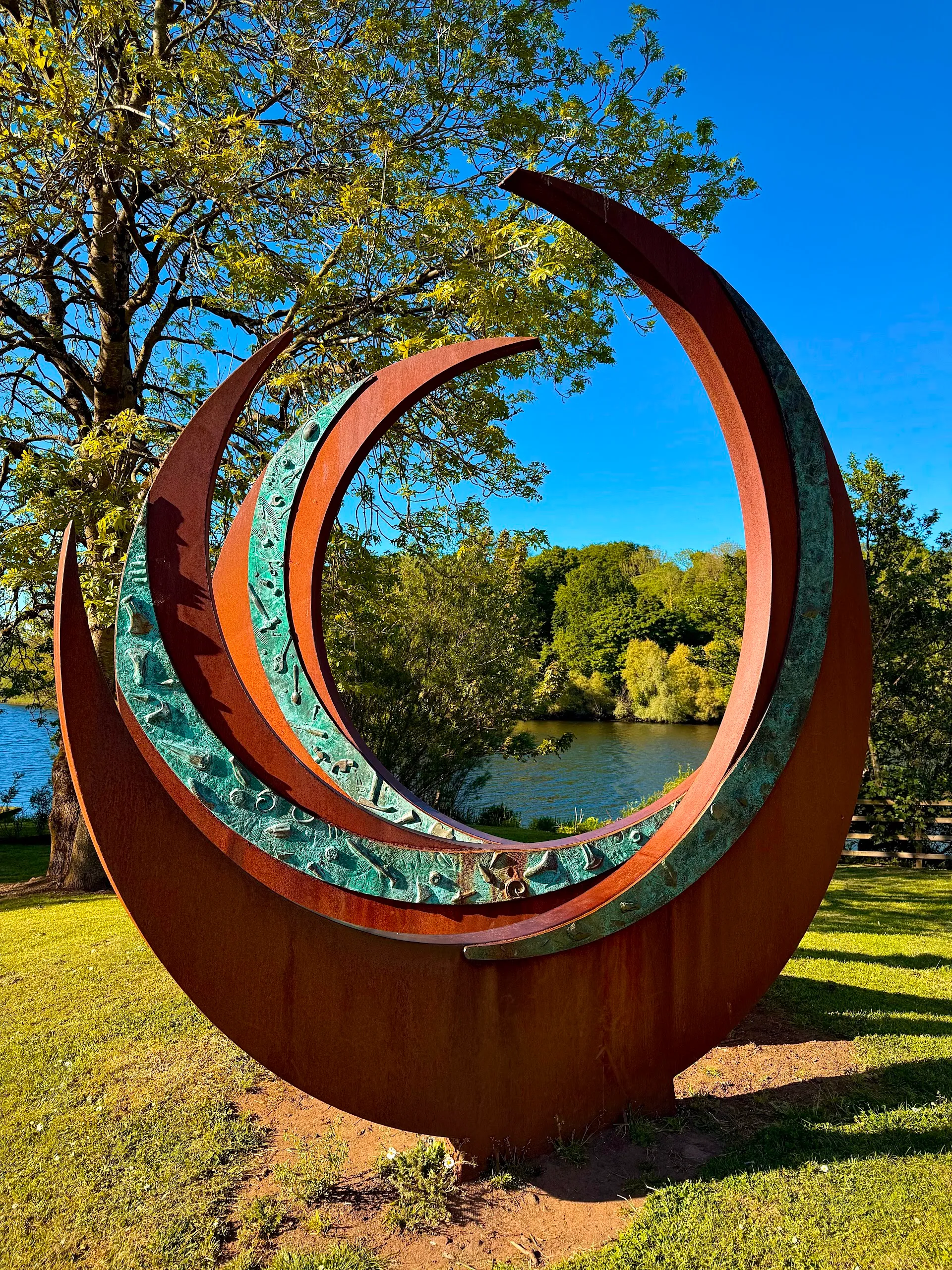 Modern crescent-shaped metal sculpture in Riverside Park, Ballymoney, with a backdrop of trees and the River Bann.