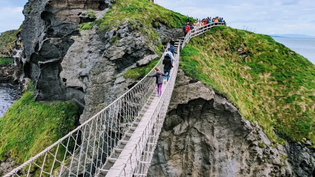 Tourists crossing the Carrick-a-Rede Rope Bridge in Northern Ireland, suspended between cliffs with views of the rugged coastline.