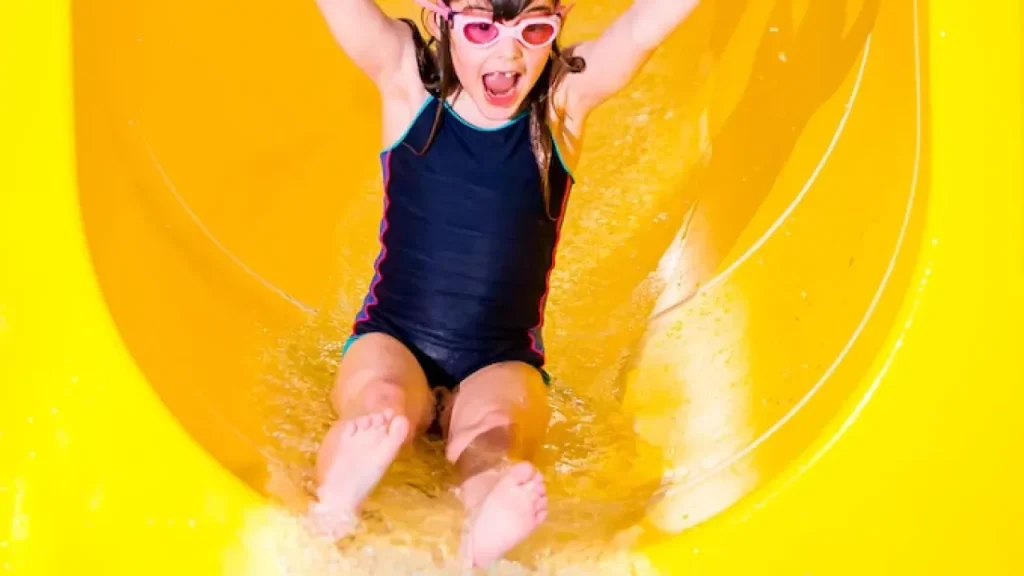Excited young girl with pink goggles and arms raised in joy sliding down bright yellow curved water flume at Perth leisure pool, demonstrating the thrilling family attractions