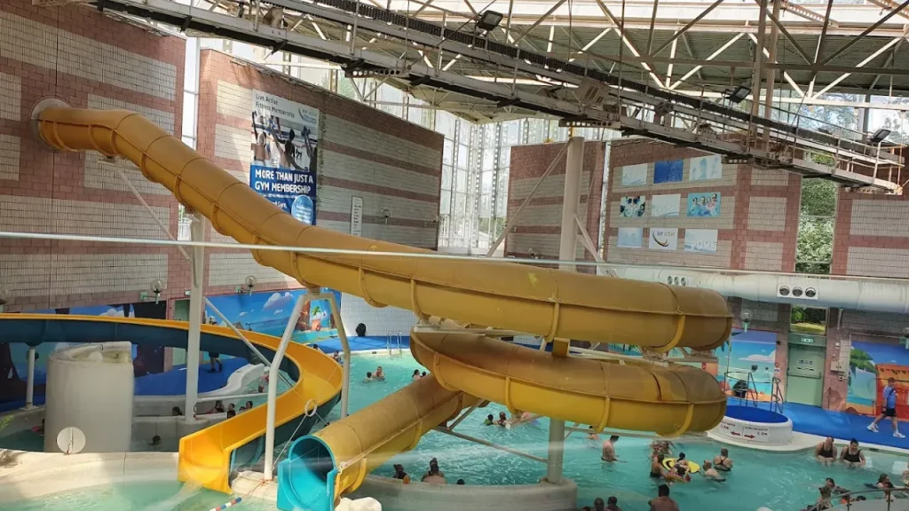Wide interior view of Perth leisure pool showing large yellow curved water flumes, multiple swimming pool areas with families, brick architecture, high ceiling with natural lighting, and various water play features