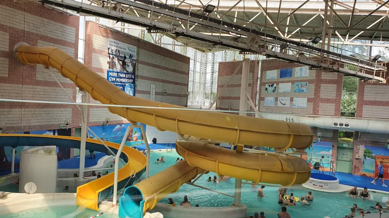 Wide interior view of Perth leisure pool showing large yellow curved water flumes, multiple swimming pool areas with families, brick architecture, high ceiling with natural lighting, and various water play features