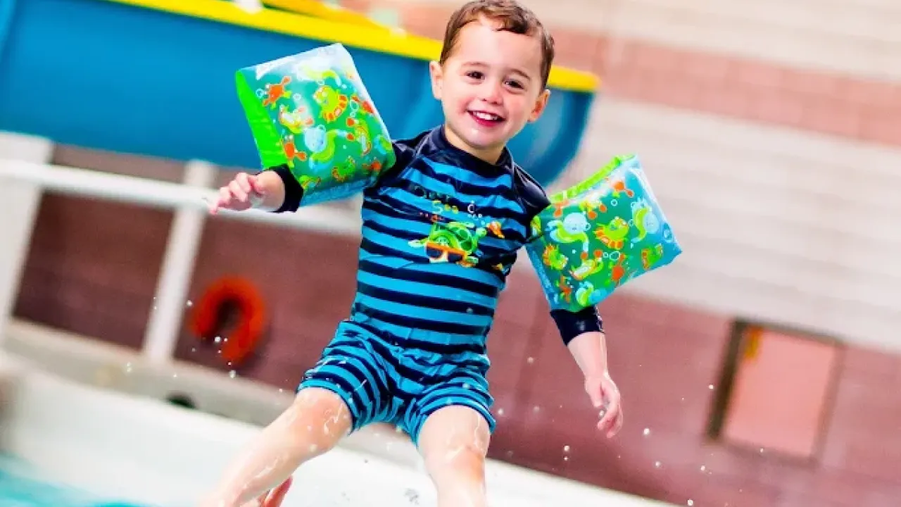 Smiling young child wearing colorful arm floats and blue striped swimsuit sitting at the edge of Perth leisure pool with feet in water, demonstrating the safe family swimming environment