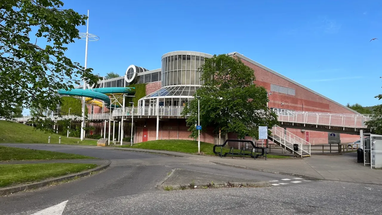 Exterior view of Perth leisure pool building showing modern brick architecture, curved glass frontage, outdoor green water slide, entrance stairs, parking area and landscaped grounds in Perth Scotland