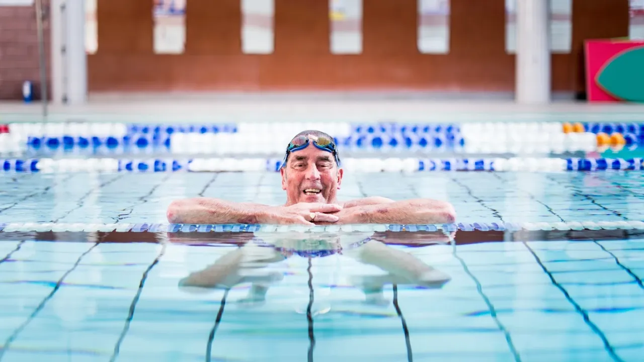 Smiling swimmer wearing goggles in Perth leisure pool's lane swimming area with clear pool lanes, lane ropes, and brick architecture visible in background