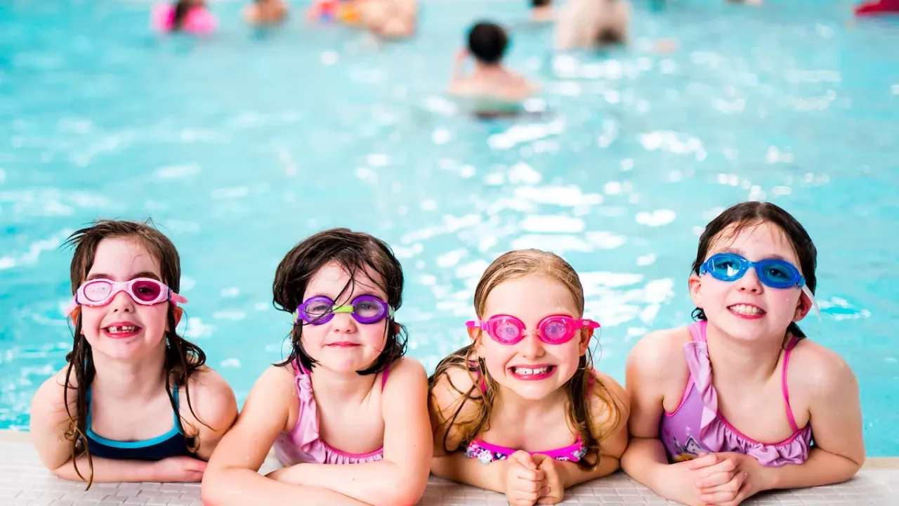 Four happy children wearing colorful swimming goggles and swimsuits posing at the edge of Perth leisure pool during swim lessons, showing the family-friendly swimming facilities