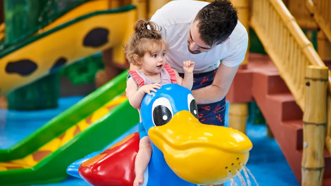 Father supervising young daughter playing on colorful pelican-shaped water toy in Perth leisure pool's children's play area, showing the safe family-friendly interactive water features