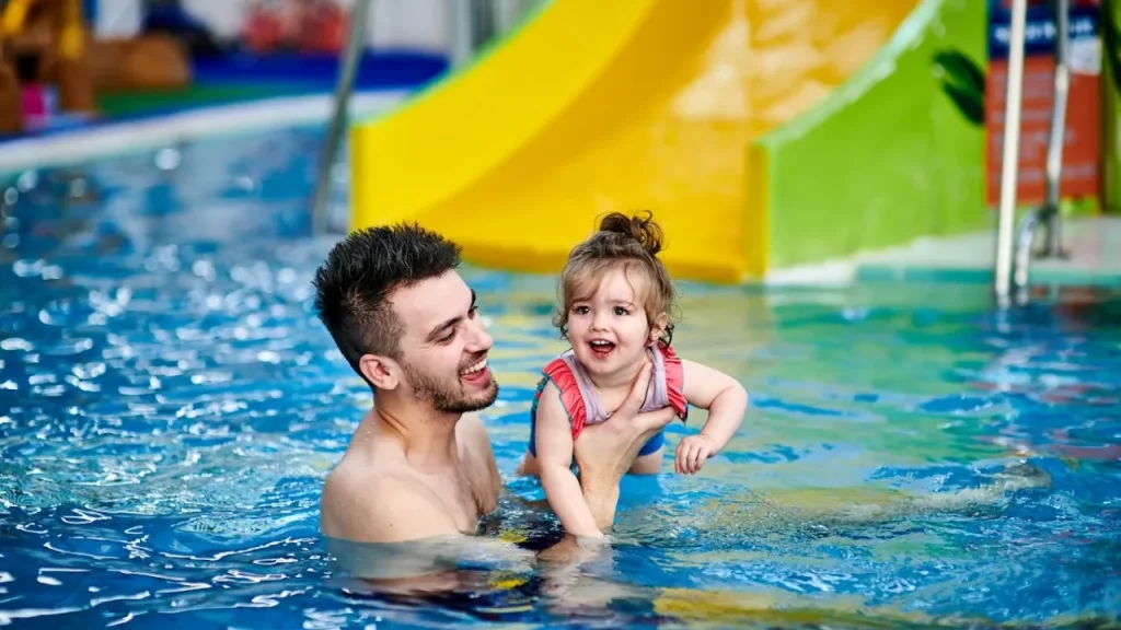 Smiling father and young daughter with water wings enjoying together in Perth leisure pool's shallow children's area with colorful water slides visible in background, demonstrating safe family swimming
