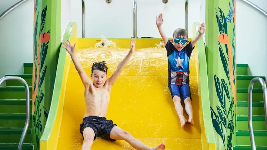Two excited children with arms raised sliding down a bright yellow water flume with tropical decorations at Perth leisure pool, showing the family-friendly slide attractions